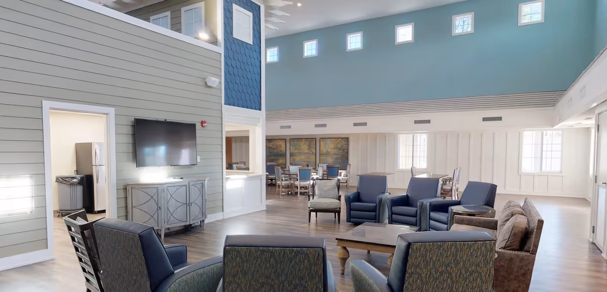 A spacious and well-lit common area in a senior living facility featuring multiple seating options including armchairs and sofas arranged around coffee tables. The room has high ceilings with small square windows near the top, light blue and gray walls, and wood flooring. A flat-screen TV is mounted on the wall above a decorative cabinet, and a dining area with tables and chairs is visible in the background.