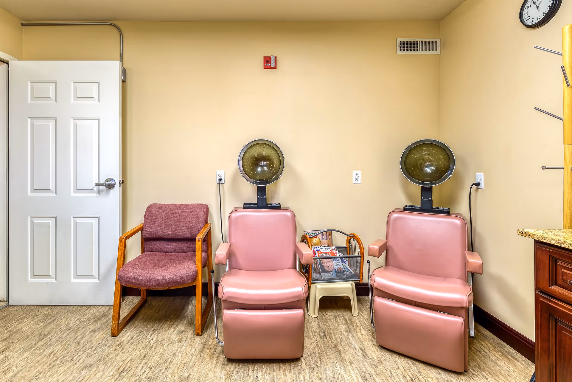Interior room with two vintage pink salon hair dryers, a maroon upholstered chair, a small table with magazines, a white door, and a wall clock.