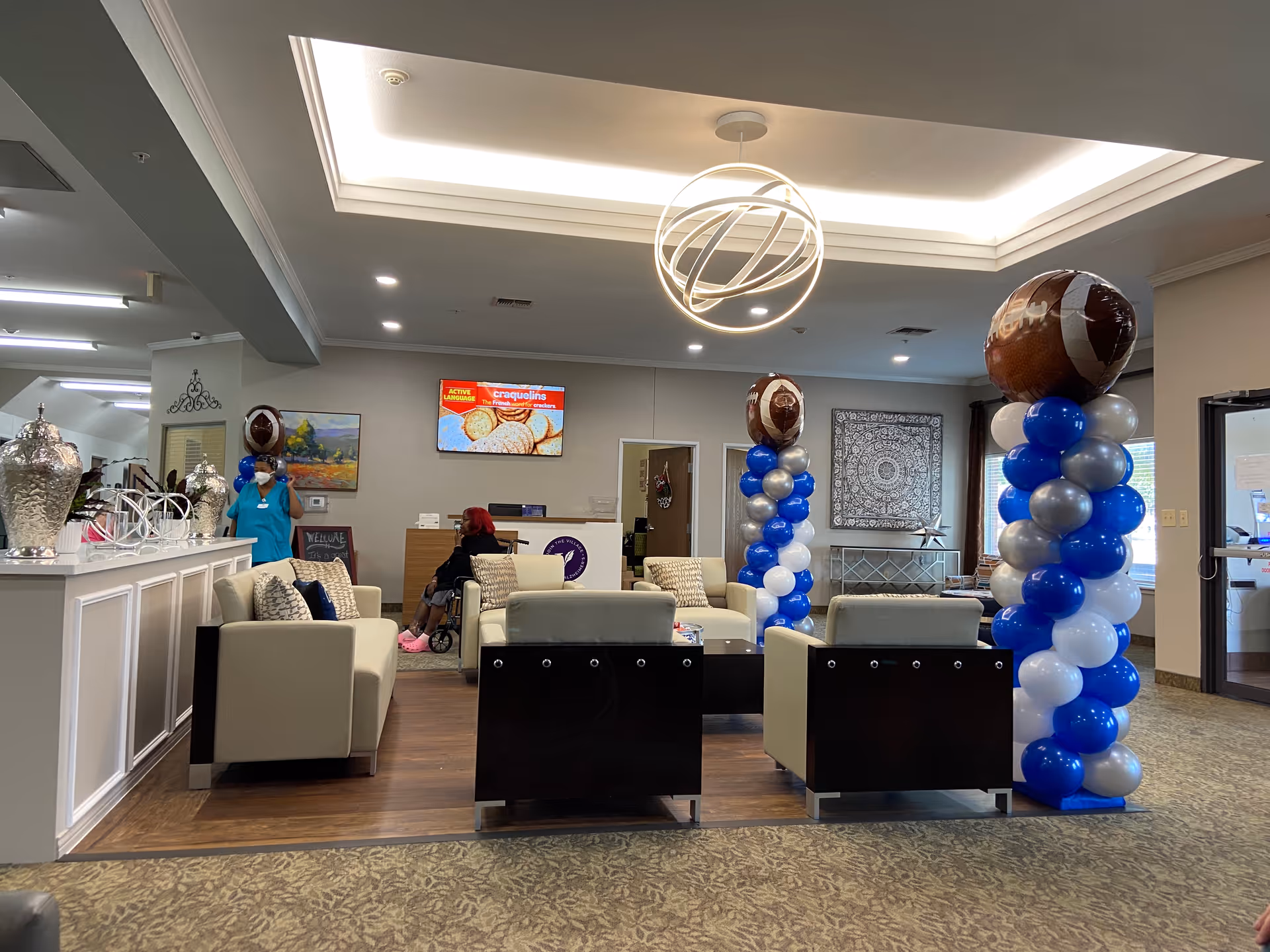 A spacious and well-lit senior living facility lobby with modern white and black armchairs arranged around a coffee table. Two tall balloon columns decorated with blue, white, and silver balloons topped with football-shaped balloons stand on either side. A woman in a wheelchair and a staff member wearing a mask are visible in the background near the reception desk. The ceiling features a stylish circular light fixture, and there is a TV mounted on the wall displaying an advertisement. The room has a warm and welcoming atmosphere with decorative elements including a painting and a patterned wall hanging.