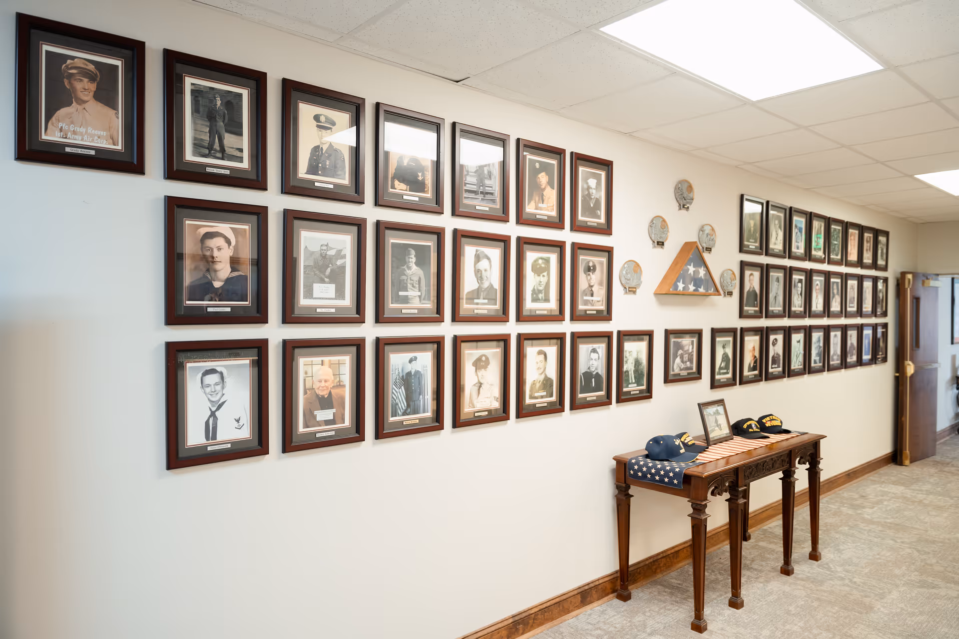 A hallway wall decorated with numerous framed black and white and color photographs of military veterans, arranged in a grid. Below the photos is a wooden table displaying folded American flags and several military hats. The hallway has a carpeted floor and a drop ceiling with fluorescent lighting.