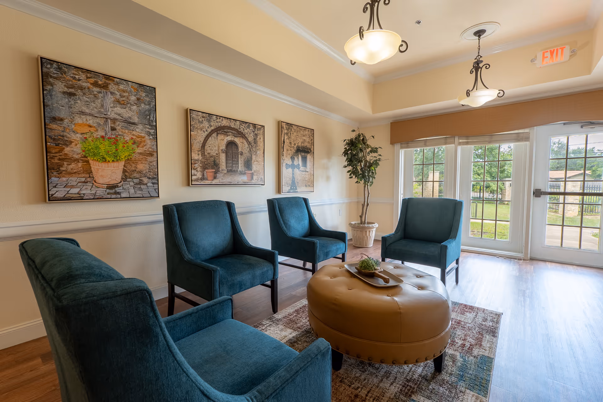 A cozy seating area in a senior living facility with four teal upholstered armchairs arranged around a large round tan ottoman on a patterned rug. The room has light-colored walls adorned with three framed pictures of rustic stone scenes and a potted plant in the corner near large windows and a glass door letting in natural light.