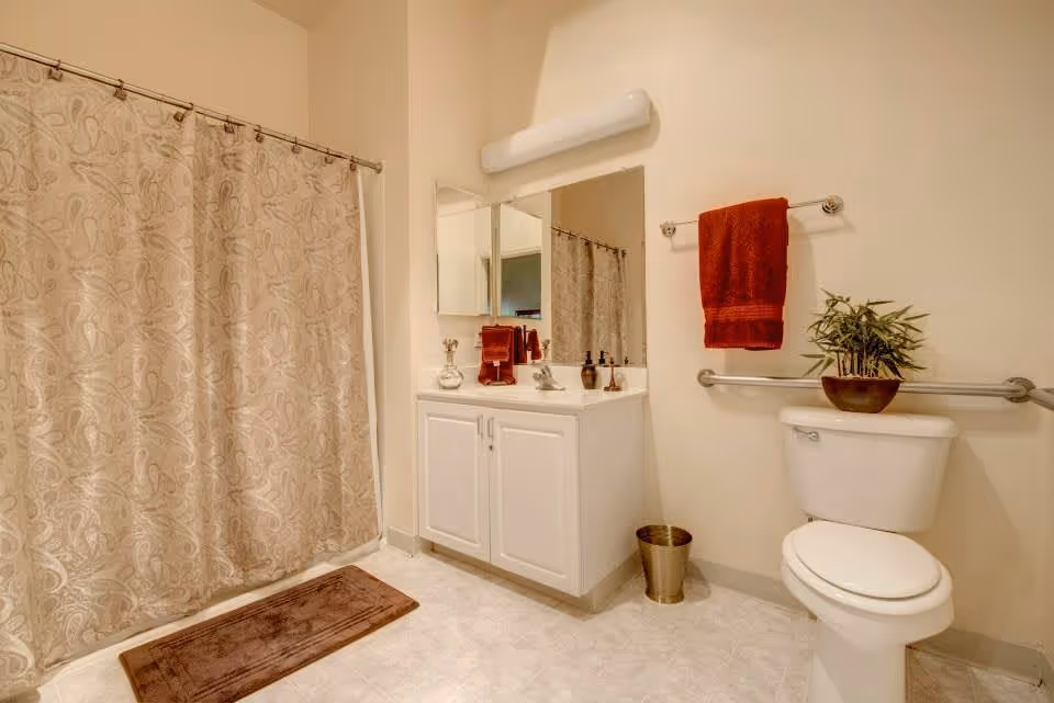 A clean bathroom featuring a white toilet with a potted plant on the tank, a towel rack with a red towel, a white vanity with a sink, a large mirror above the sink, and a shower with a beige patterned curtain. There is also a brown bath mat on the tiled floor and a small trash can next to the vanity.