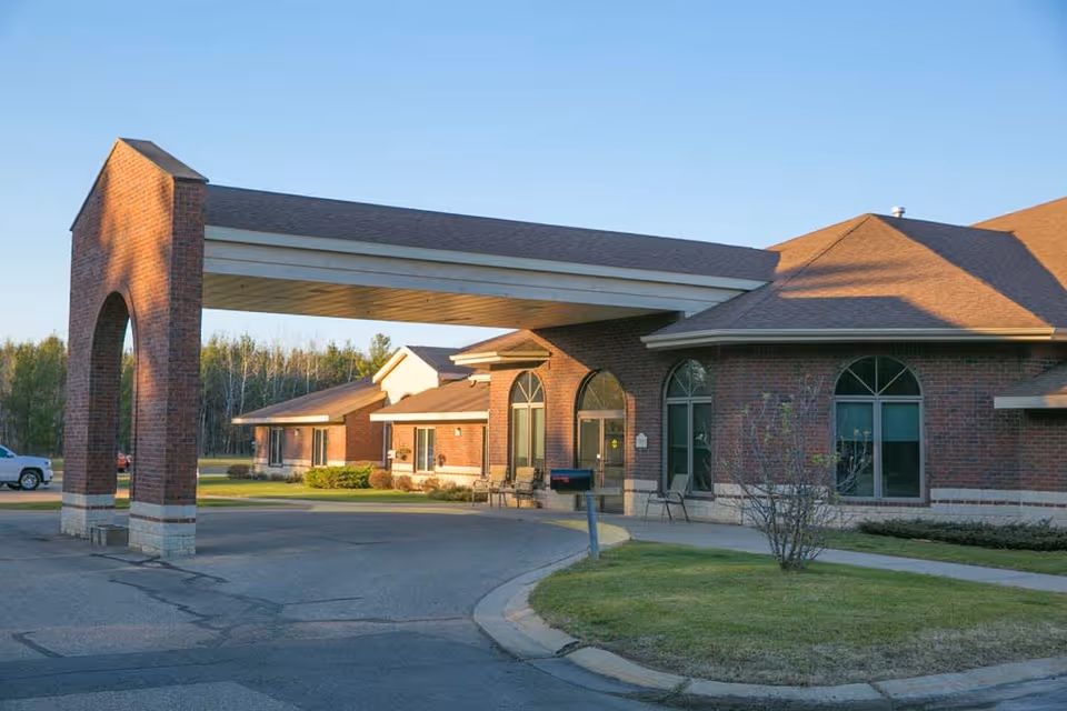 Exterior view of a senior living facility with a covered entrance supported by brick pillars. The building has large arched windows and a well-maintained lawn with a few small bushes. A white vehicle is parked in the background under a clear blue sky.