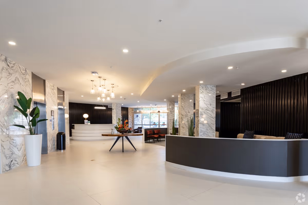 Bright modern lobby with reception desks, seating area, a central table with flowers, marble-accented columns and plants.