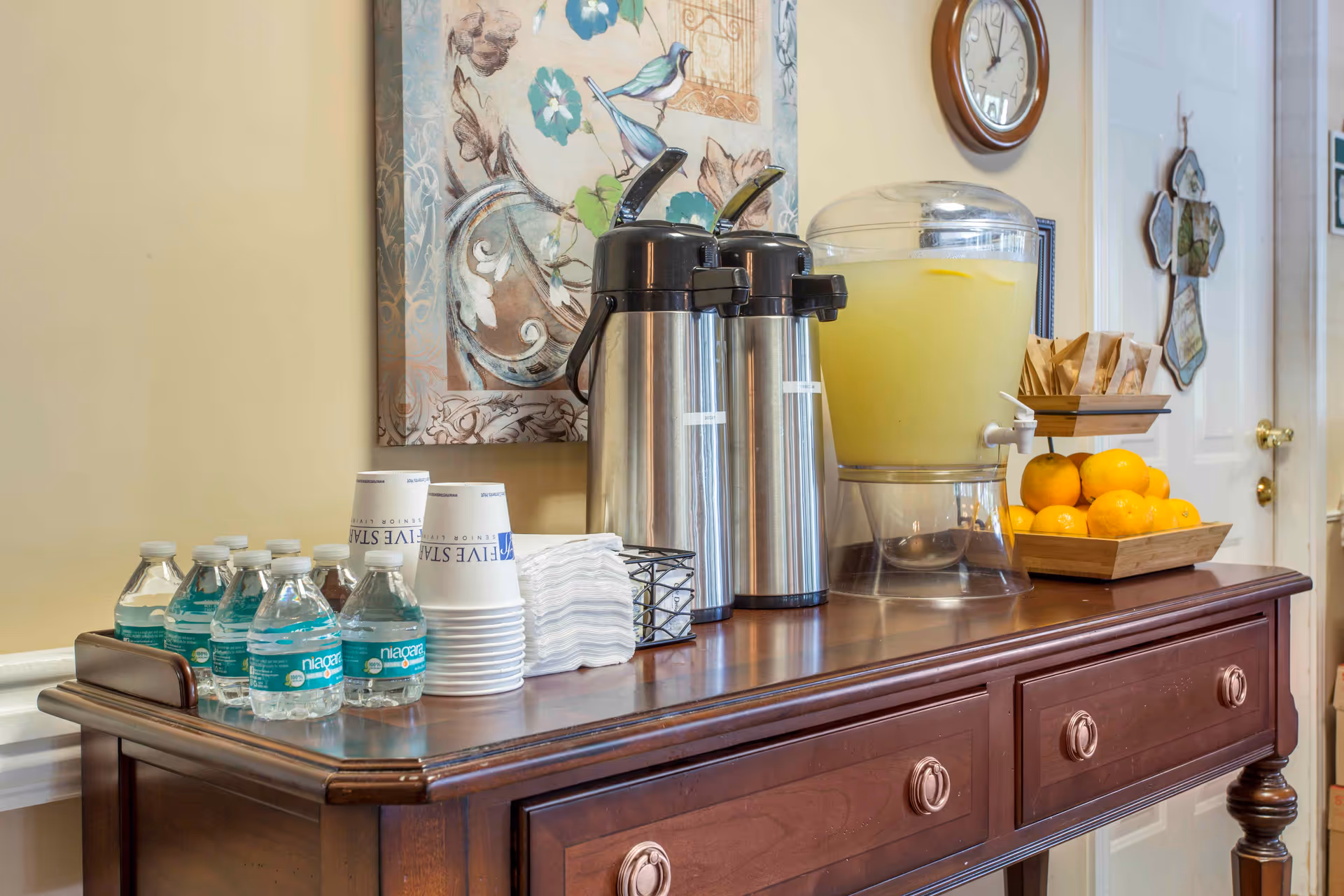 A wooden sideboard table with two stainless steel airpots, a large clear beverage dispenser filled with lemonade, several bottles of water, stacked disposable cups, napkins, a small basket with stirrers, a tray of lemons, and a wall clock above. A decorative painting with birds and flowers hangs on the wall behind the table.