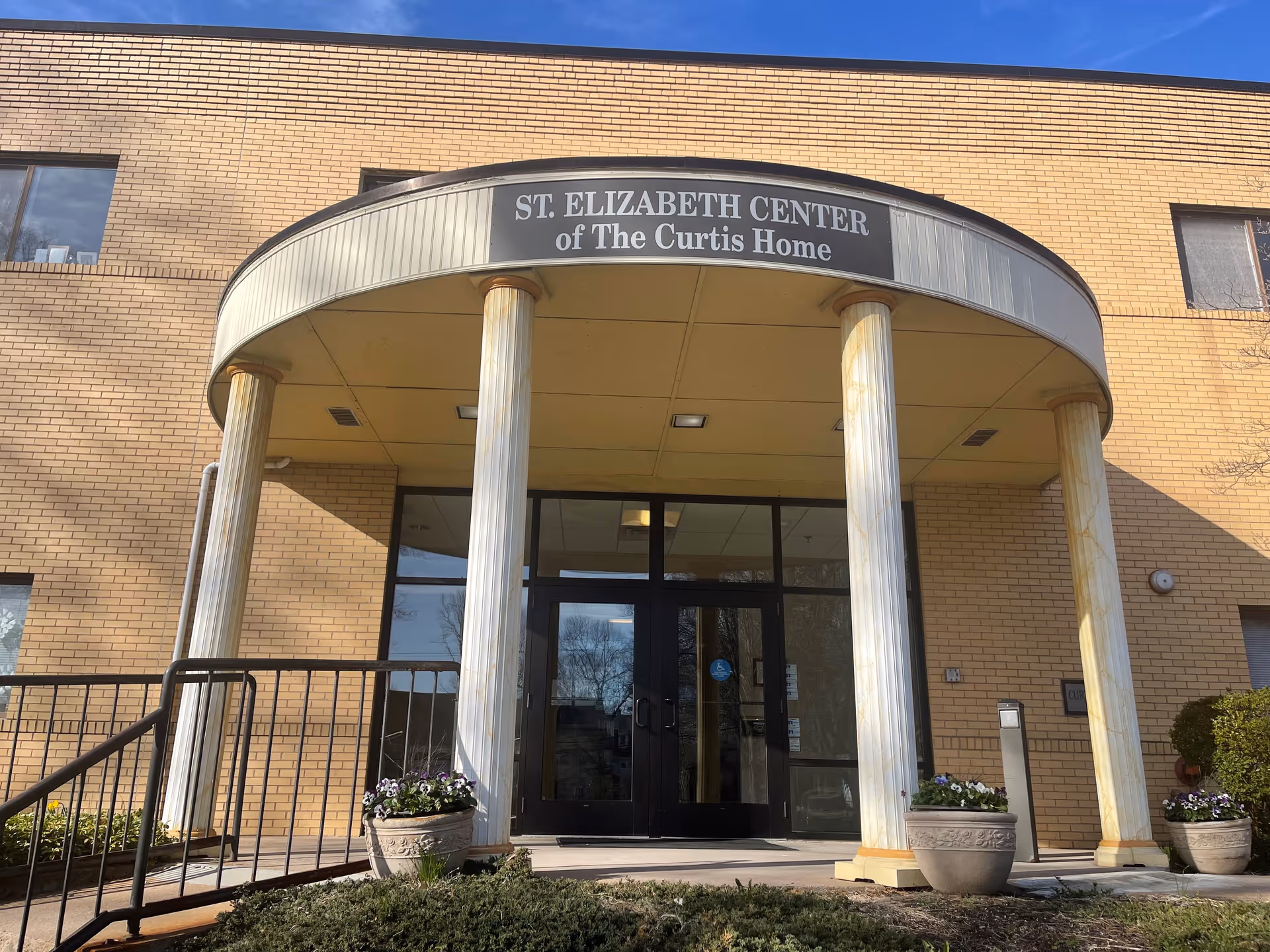 Entrance of the St. Elizabeth Center of The Curtis Home, featuring a curved portico supported by four white columns, with a brick building facade and glass double doors under a clear blue sky. There are potted plants on either side of the entrance and a railing along the steps.