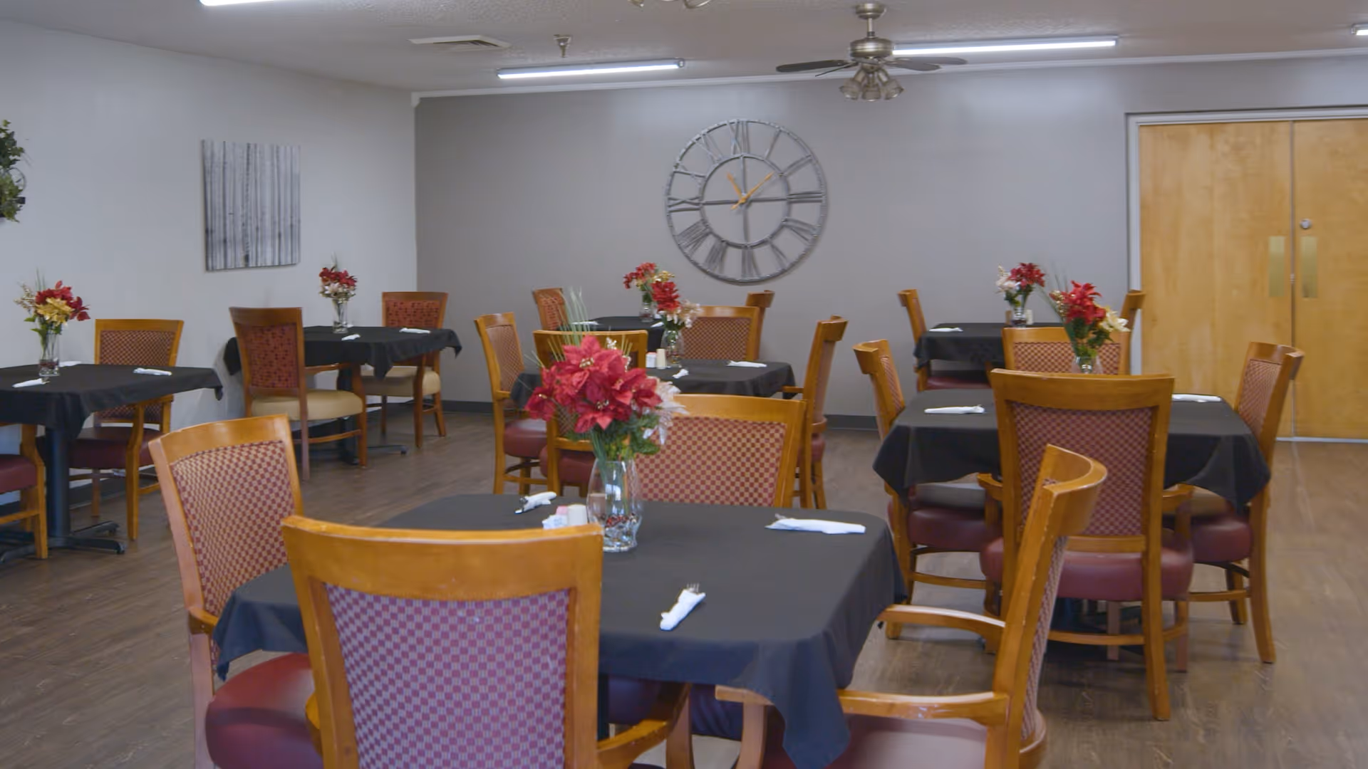 Dining room with multiple wooden tables covered with black tablecloths, each set with napkins and decorated with vases of red flowers. Wooden chairs with red cushioned seats surround the tables. A large wall clock and a ceiling fan are visible on the light gray walls.