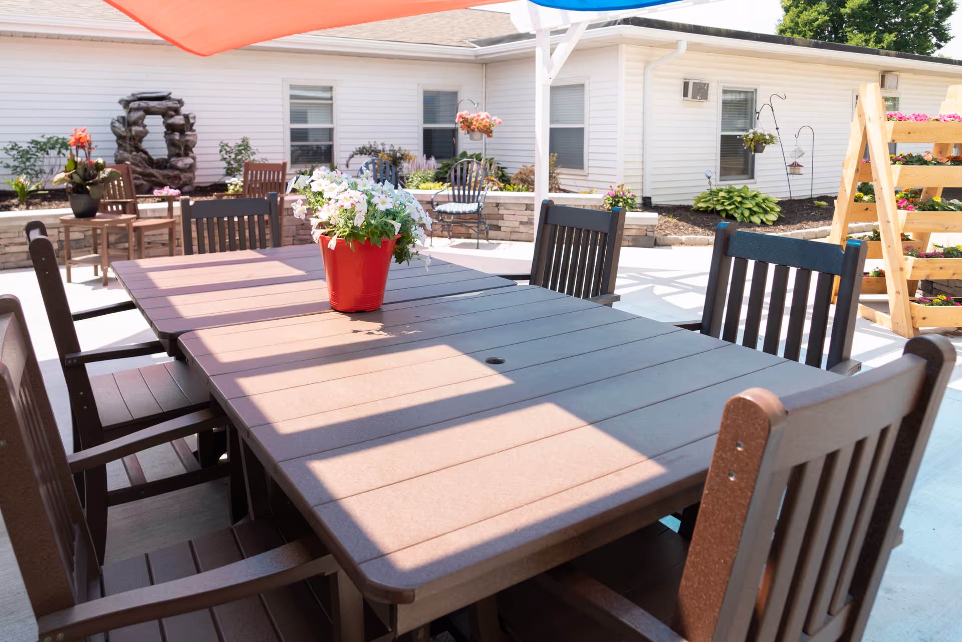 Outdoor patio area with a large rectangular table surrounded by wooden chairs. A red pot with white flowers is placed in the center of the table. The patio is shaded by a large red and blue canopy. In the background, there are more chairs, plants, and a white building with windows.
