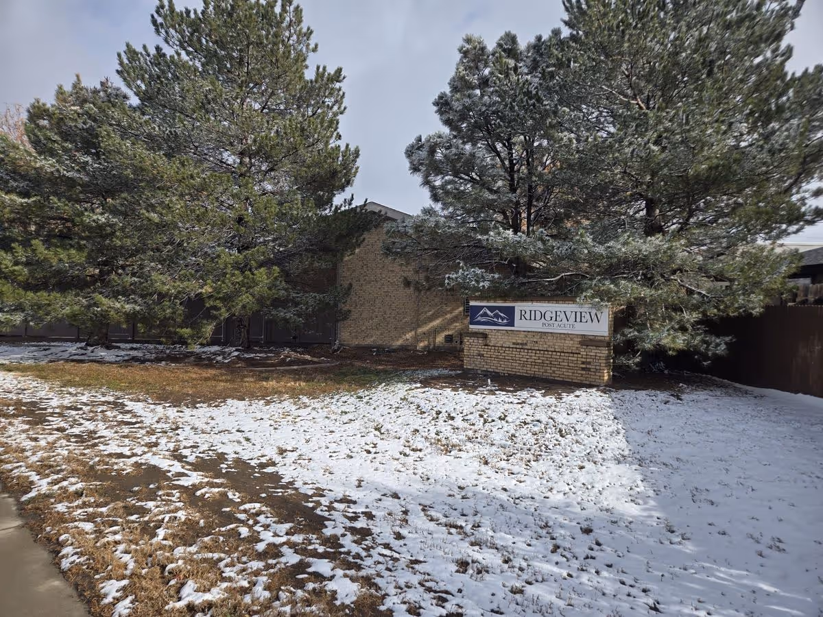 Snow-dusted lawn and pine trees in front of a brick building with a 'Ridgeview Post Acute' sign.