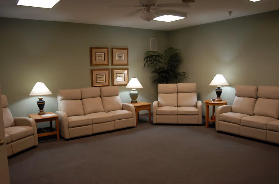 A cozy living room with four beige recliner sofas arranged in a semi-circle. There are three wooden side tables, each with a lamp on top. The walls are painted light green and decorated with four framed butterfly pictures. A large potted plant is placed in the corner of the room. The ceiling has a fan and a fluorescent light fixture.