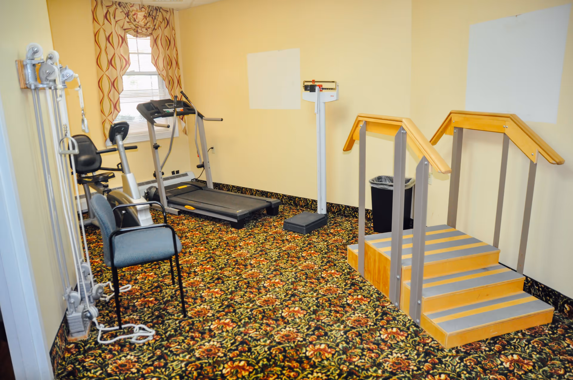 A small exercise room with patterned carpet, featuring a treadmill, stationary bike, weight machine, a chair, a scale, and a set of wooden stairs with handrails for physical therapy or rehabilitation exercises. The room has light yellow walls and a window with patterned curtains.