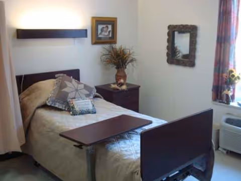 A furnished bedroom in a nursing facility with a bed, overbed table, nightstand, and window.