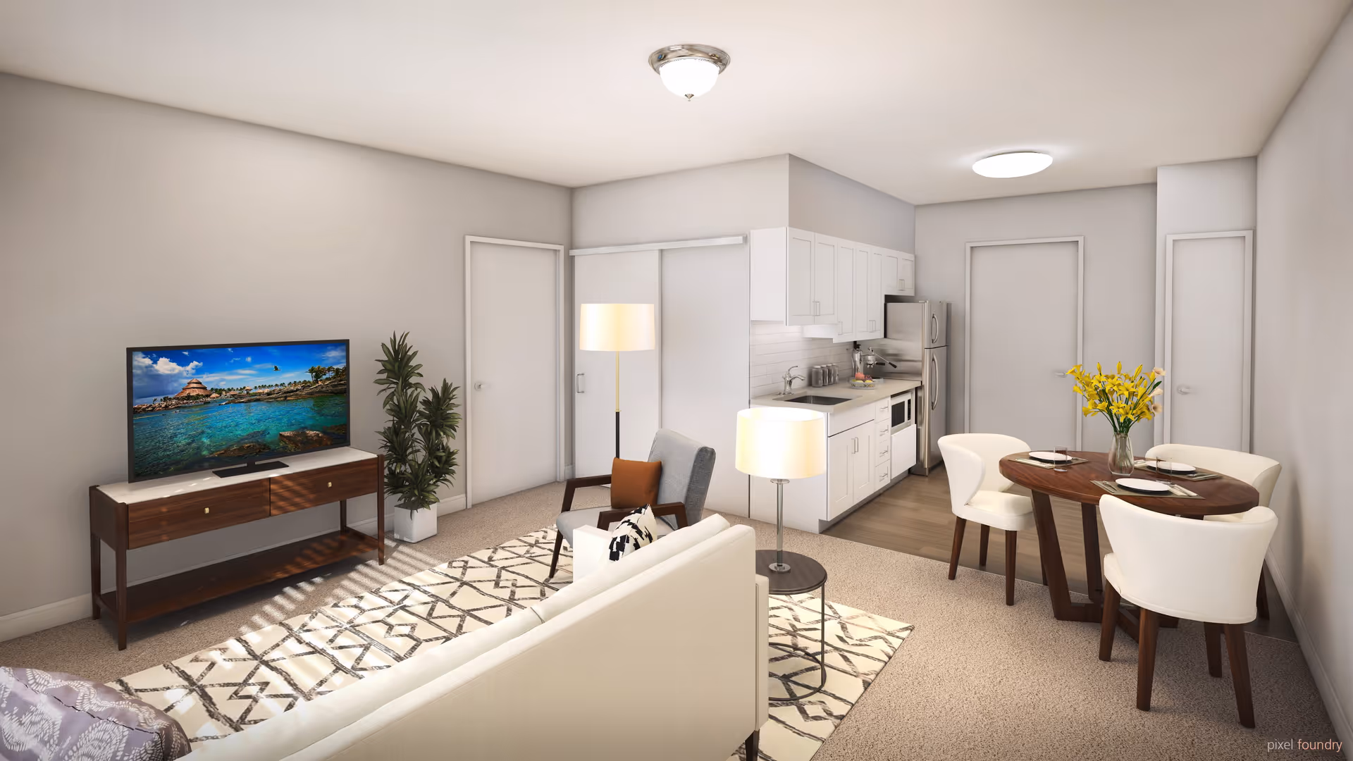 A modern, open-concept living space in an assisted living facility featuring a white sofa with pillows, a patterned rug, a wooden TV stand with a flat-screen TV displaying a scenic ocean view, a potted plant, a small round dining table with four white chairs and a vase of yellow flowers, and a compact kitchen area with white cabinets, stainless steel appliances, and a countertop with a sink and microwave.