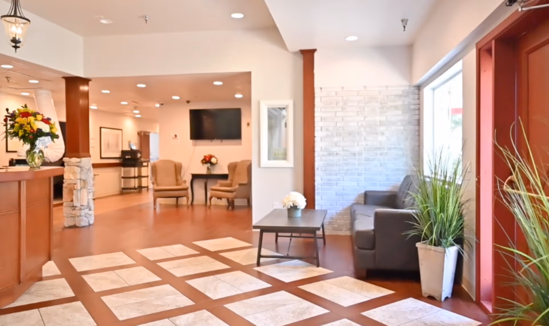 A bright and welcoming healthcare center lobby with a wooden reception desk on the left adorned with a vase of flowers. The floor features a patterned tile design leading to a seating area with a gray sofa, a coffee table with a small flower arrangement, and two beige armchairs near a wall-mounted TV. Potted plants are placed near the large window and entrance door on the right.
