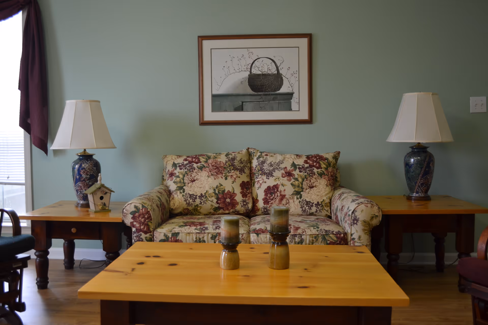 A cozy living room with a floral patterned loveseat centered against a light green wall. On either side of the loveseat are wooden side tables, each with a blue ceramic lamp with a white lampshade. A framed picture of a basket hangs above the loveseat. In front of the loveseat is a wooden coffee table with two candles on it. A window with a dark curtain is partially visible on the left side.