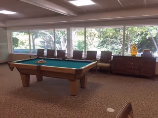 A spacious room with a pool table in the center, several chairs lined up against large windows that show a view of trees outside, and a wooden cabinet with decorative items on top.