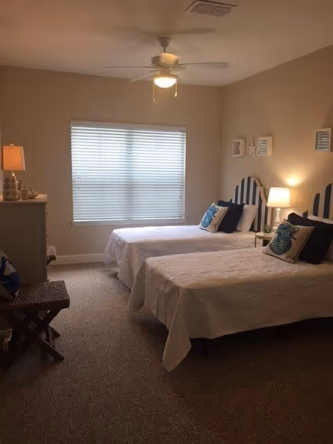 A bedroom with two twin beds featuring blue and white striped headboards. Each bed has white bedding and decorative pillows. There are two bedside tables with lamps, a ceiling fan with a light, a window with blinds, a dresser with a lamp, and a small bench at the foot of one bed. The walls are painted beige and the carpet is light brown.