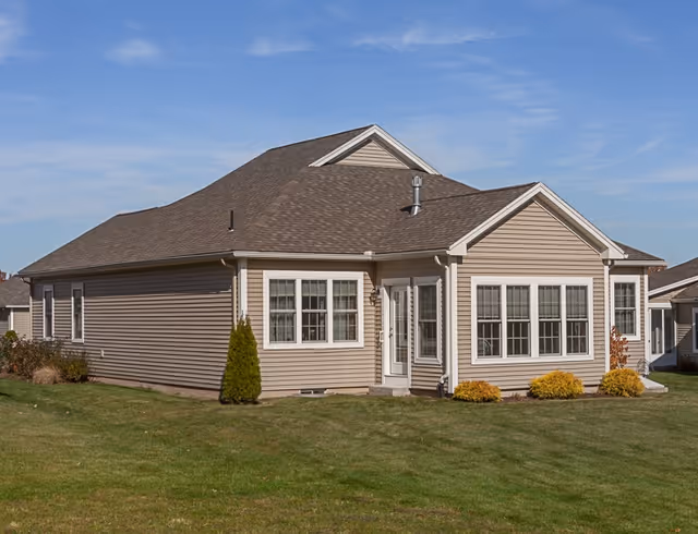 Exterior view of a single-story beige house with multiple windows and a brown shingled roof, surrounded by a green lawn and some small shrubs under a partly cloudy sky.