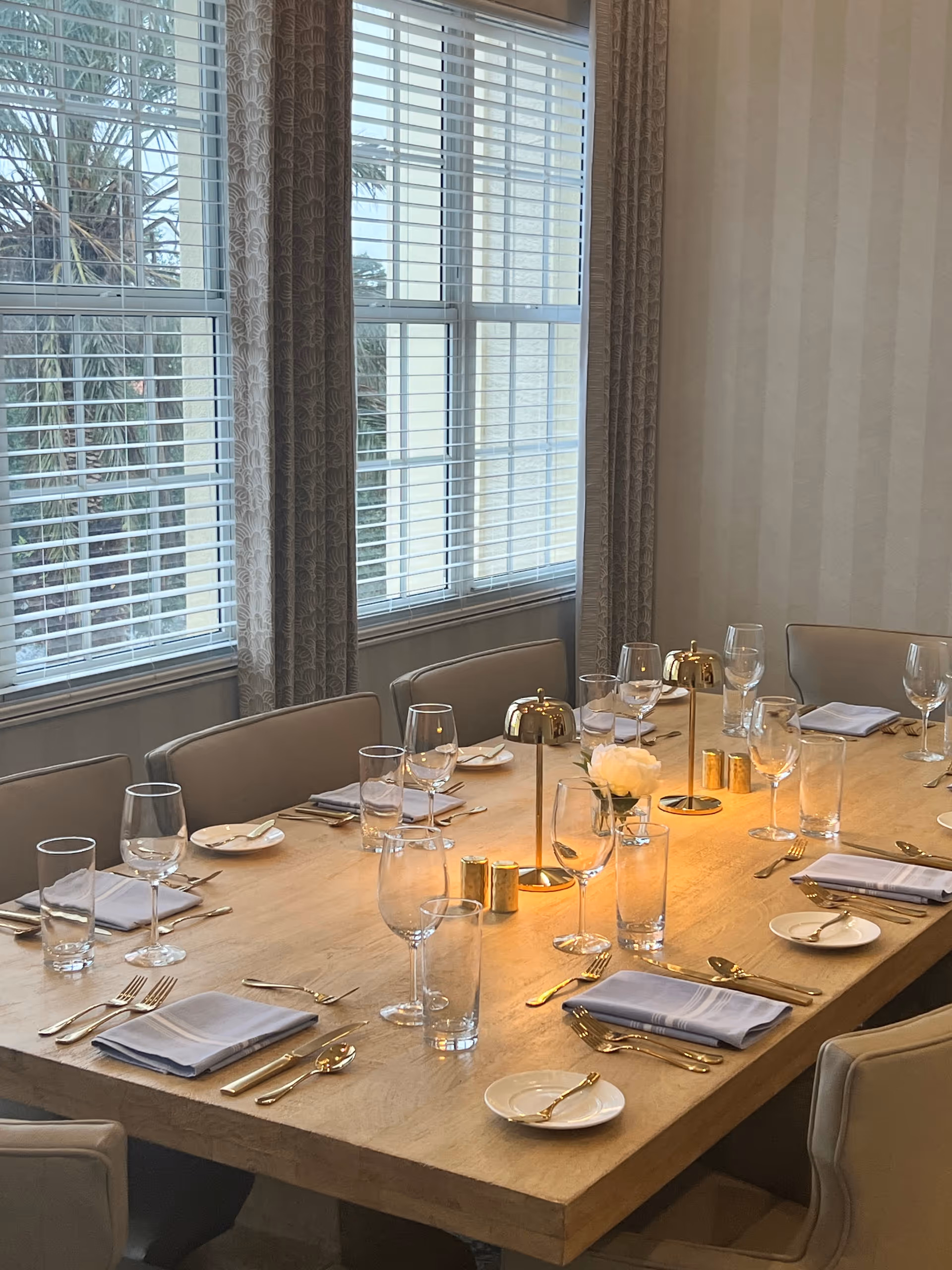 A dining table set for a meal with beige upholstered chairs around it. The table has gold-colored cutlery, white napkins, clear drinking glasses, wine glasses, small white plates, and gold candle holders. There is a small white flower centerpiece. Large windows with white blinds and patterned curtains are in the background.