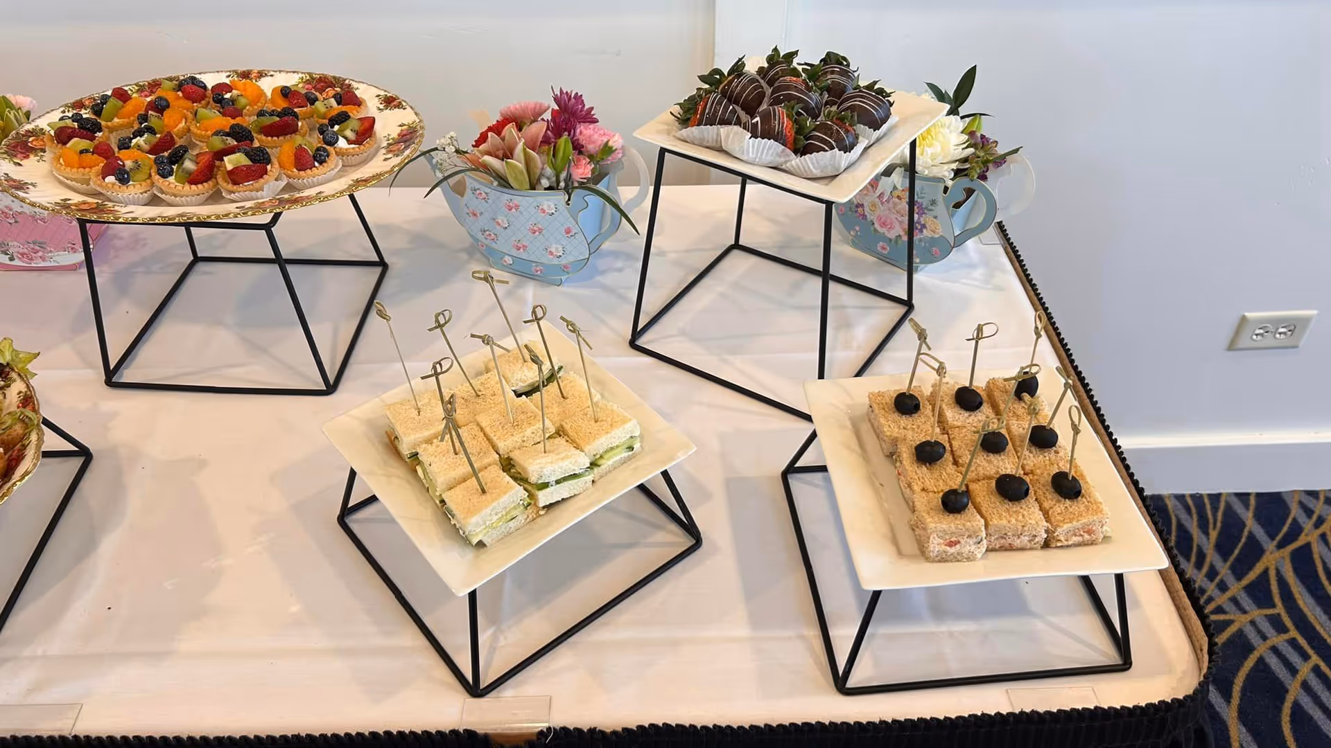 A table set with an assortment of finger foods including small sandwiches with toothpicks, fruit tarts topped with various berries, and chocolate-covered strawberries. The food is displayed on white plates elevated by black metal stands. There are also small floral arrangements in decorative teapot-shaped containers on the table.