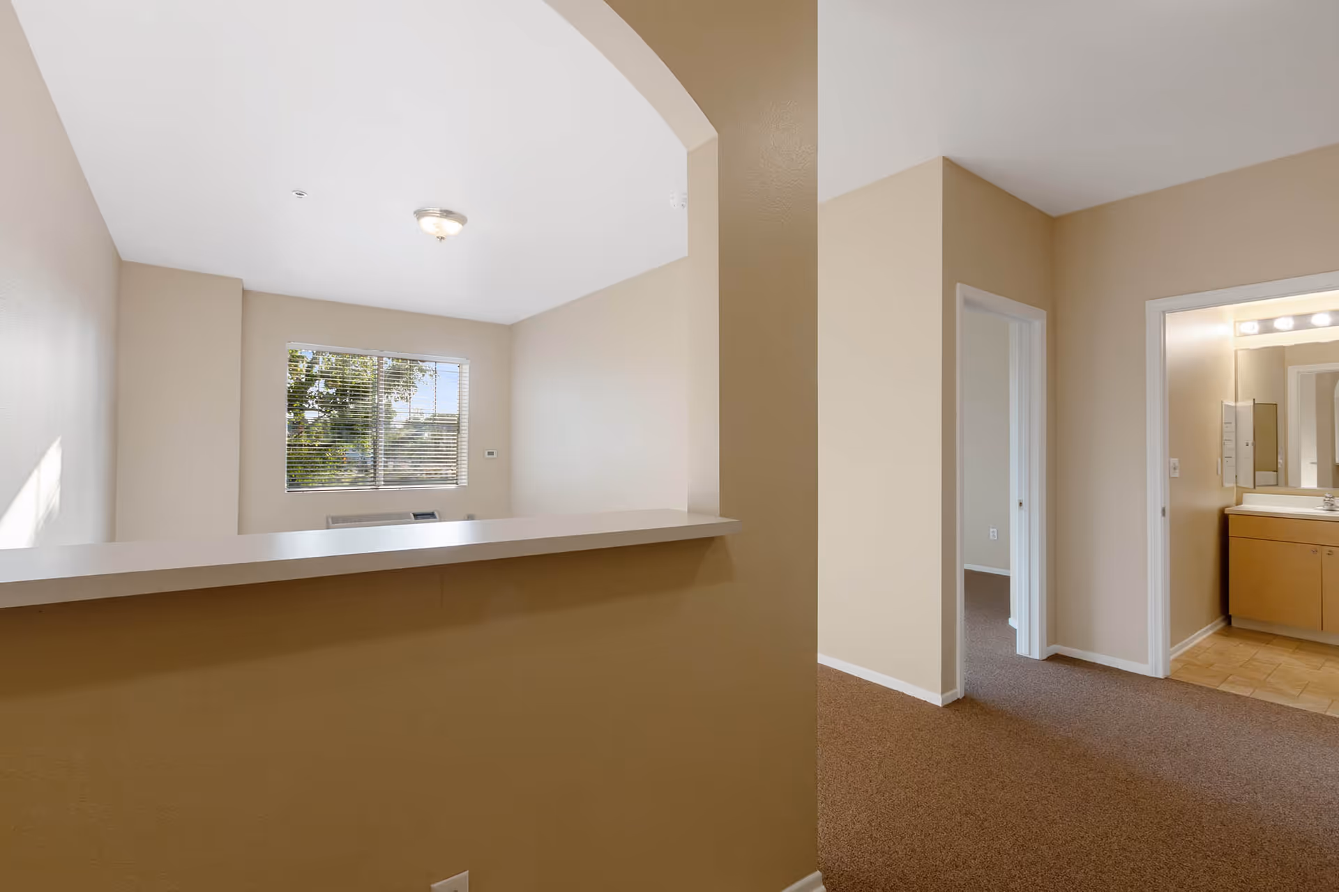 Interior view of an apartment at Claremont Place showing a beige wall with a pass-through counter, a window with blinds, a hallway leading to a bedroom and a bathroom with a vanity and mirror.