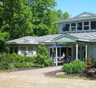 Exterior view of a single-story residential building with a porch, surrounded by greenery and trees. A person is sitting on the porch under an umbrella.
