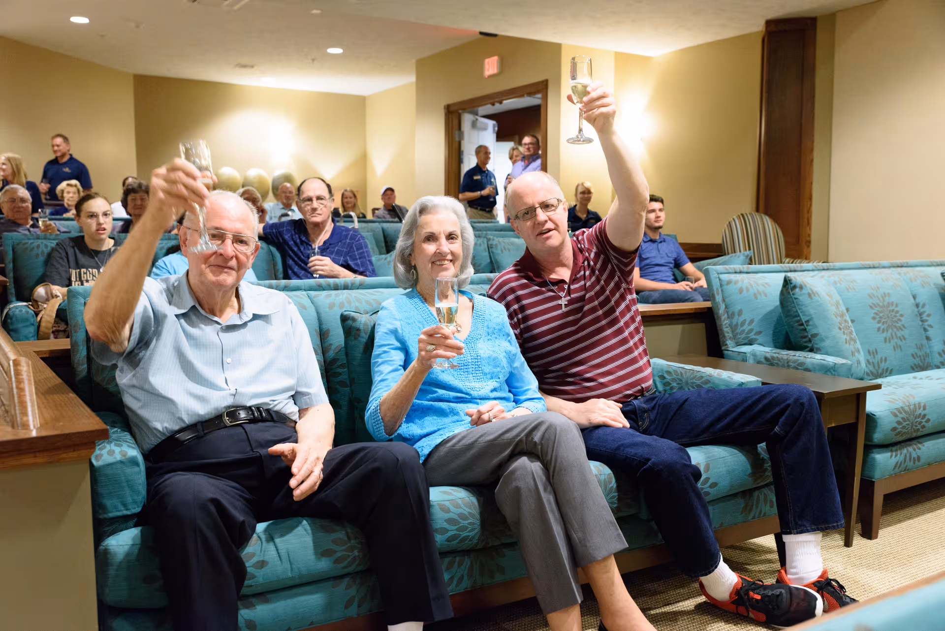 A group of elderly people sitting on teal patterned couches in a common room, with three individuals in the front raising glasses in a toast. Other residents are seated behind them, and the room has warm lighting and beige walls.