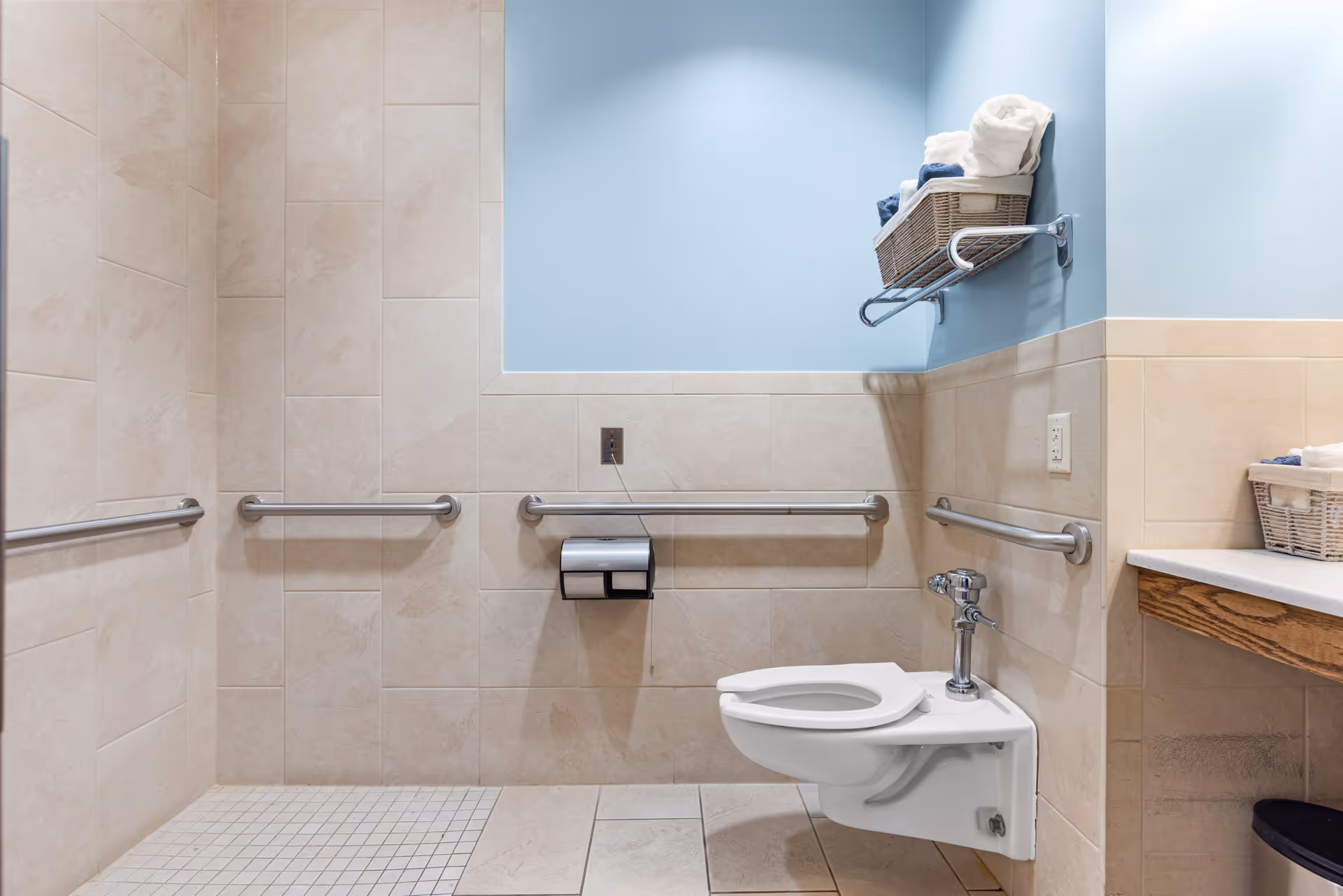 Accessible bathroom with beige tiled walls and floor, a wall-mounted toilet with grab bars on three sides, a toilet paper holder, and a shelf holding baskets with towels against a light blue painted upper wall.