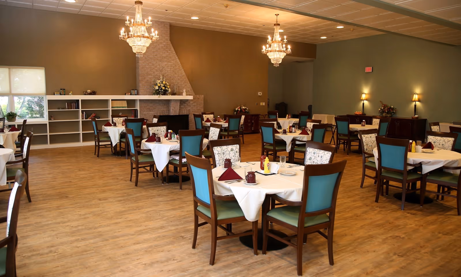 A spacious dining room with multiple round tables covered with white tablecloths, each set with maroon napkins, condiments, and glassware. The chairs have wooden frames with teal and patterned upholstery. The room features wooden flooring, a brick fireplace, chandeliers, and soft wall lighting. There are shelves with books and decorative items along one wall and large windows letting in natural light.