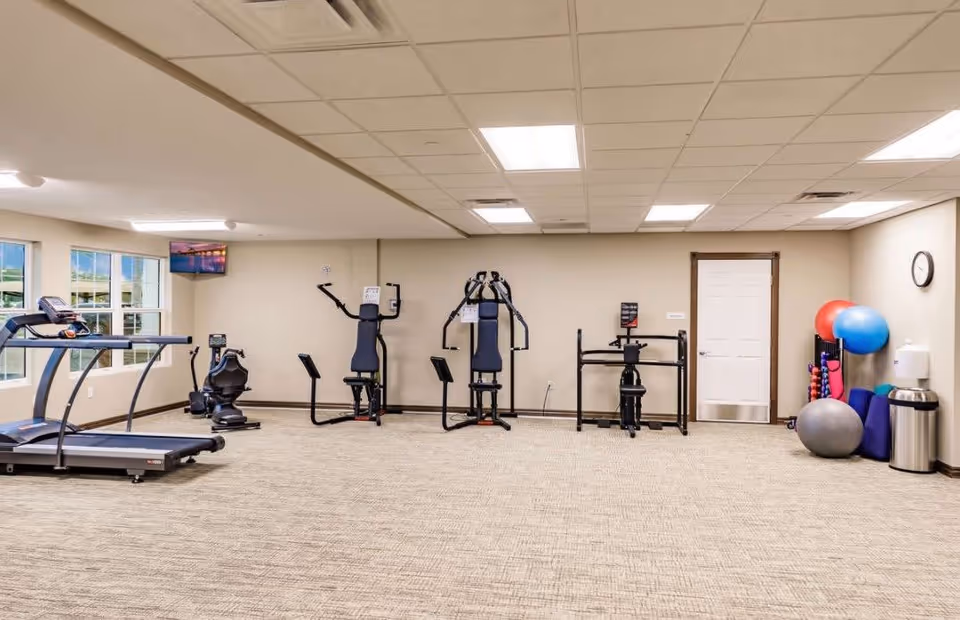 A fitness room in a senior living facility with exercise equipment including a treadmill, stationary bike, weight machines, and various exercise balls and mats neatly arranged against the wall. The room has beige walls, carpeted floor, and fluorescent ceiling lights.