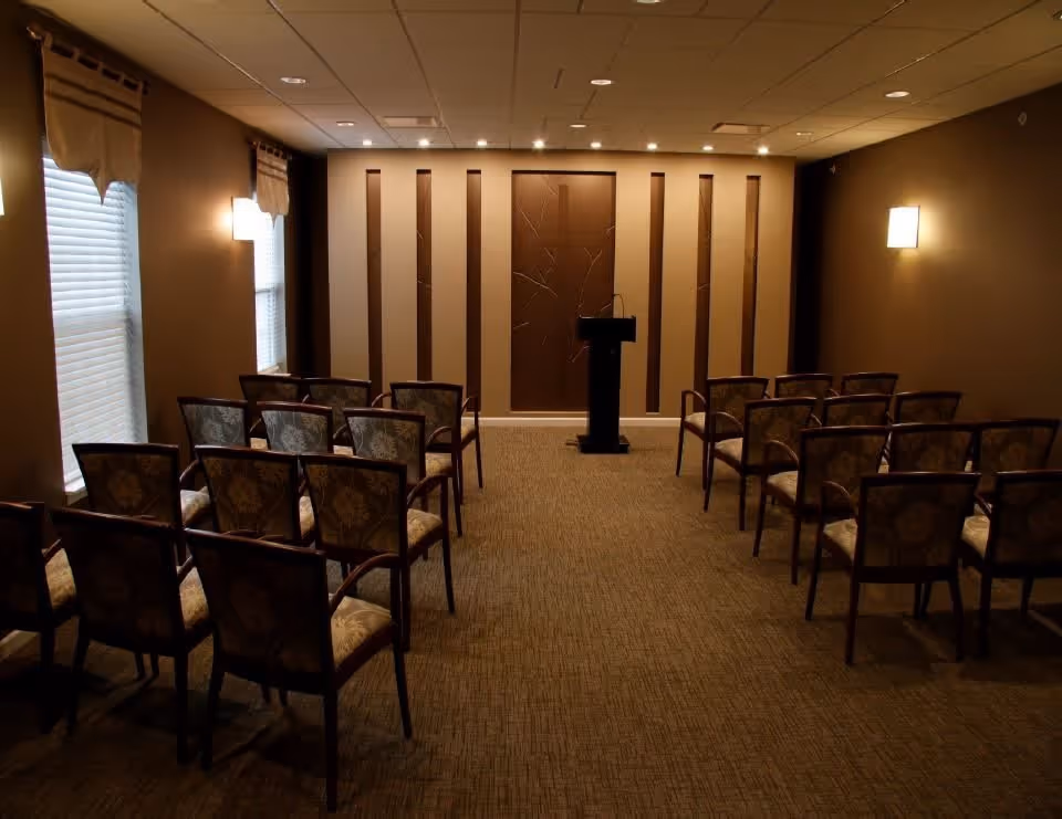 A small conference or meeting room with rows of upholstered chairs facing a podium at the front. The room has brown walls, carpeted floor, two windows with blinds and valances on the left side, and wall-mounted lights providing soft illumination.