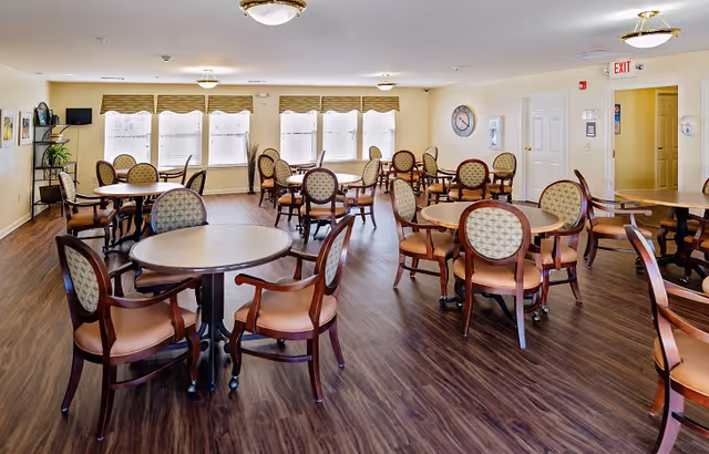A bright and spacious dining room with multiple round tables and cushioned wooden chairs arranged neatly on a wood floor. Large windows with beige valances allow natural light to fill the room. The walls are light-colored, and there is a clock and some framed artwork on the walls. Ceiling lights provide additional illumination.