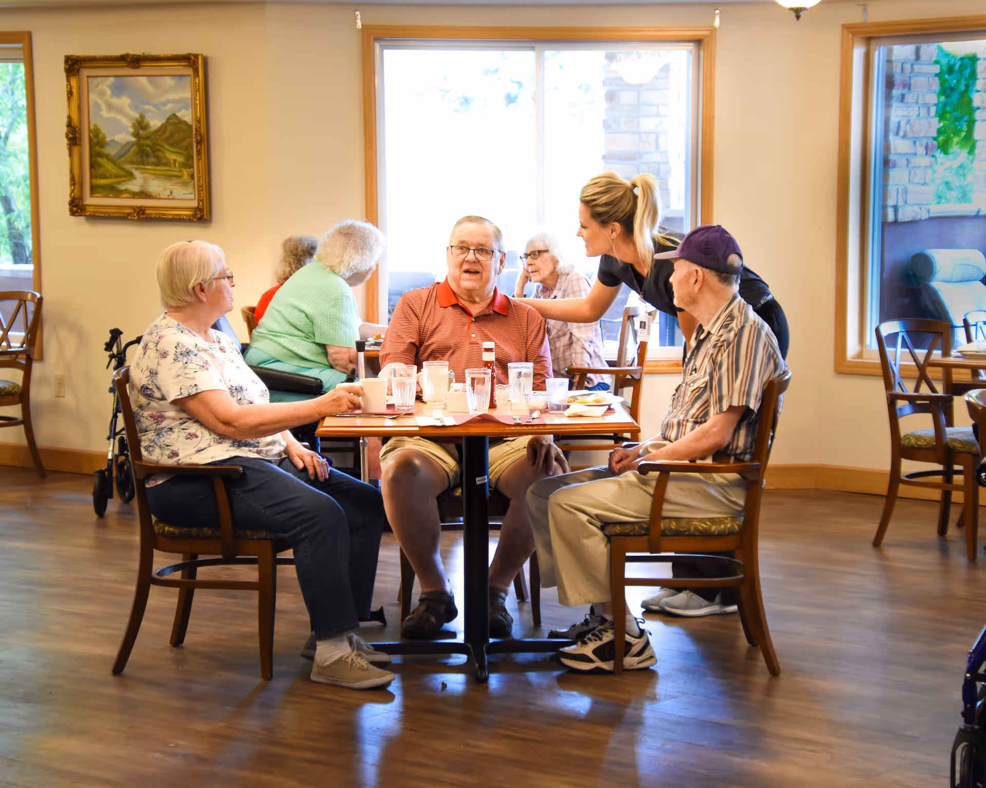 A group of elderly people sitting around a dining table in a senior living facility, with a caregiver standing and engaging with them. The room has large windows, wooden floors, and framed artwork on the wall.