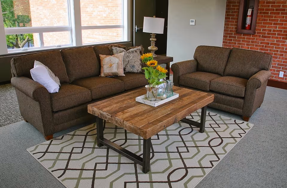 A cozy living room area with two brown upholstered sofas facing each other, a wooden coffee table with a vase of sunflowers and decorative items on top, a patterned area rug underneath, a table lamp on a side table, large windows letting in natural light, and a brick accent wall with a fire extinguisher mounted on it.
