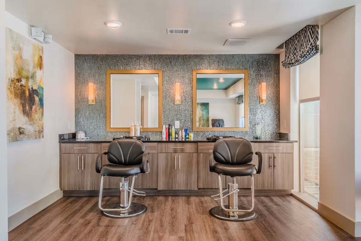 Two salon styling chairs in front of a mirrored station with wood cabinetry, decorative wall lighting, and a patterned accent wall.