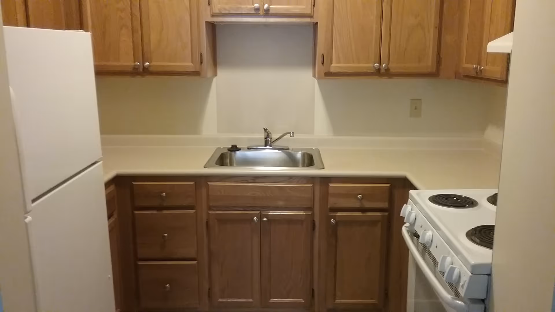 Compact kitchen with wooden cabinets, a stainless sink centered under cabinets, a white refrigerator on the left and an electric stove on the right.