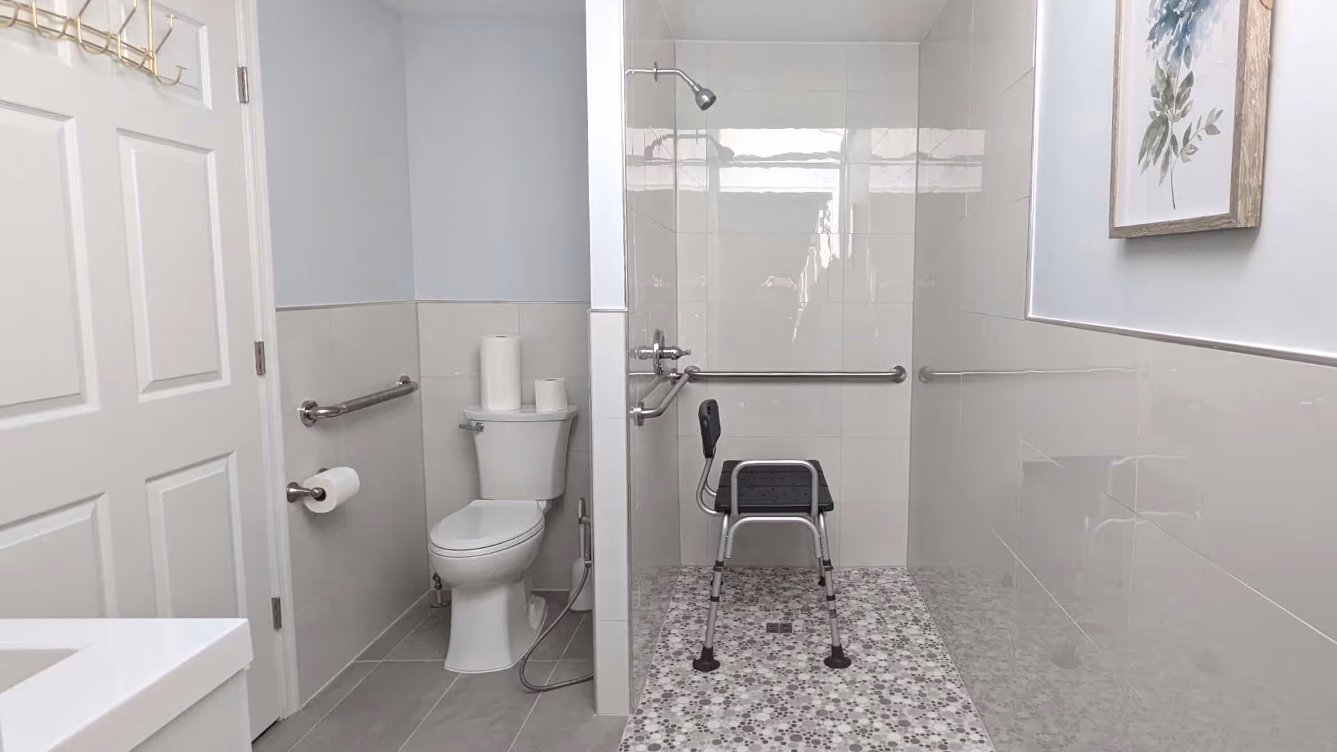 A clean and accessible bathroom featuring a white toilet with toilet paper rolls on top, a wall-mounted toilet paper holder, and grab bars on the walls. There is a walk-in shower area with a handheld showerhead and a black shower chair with a backrest. The walls are tiled halfway up with light-colored tiles, and a framed botanical print hangs on the wall.