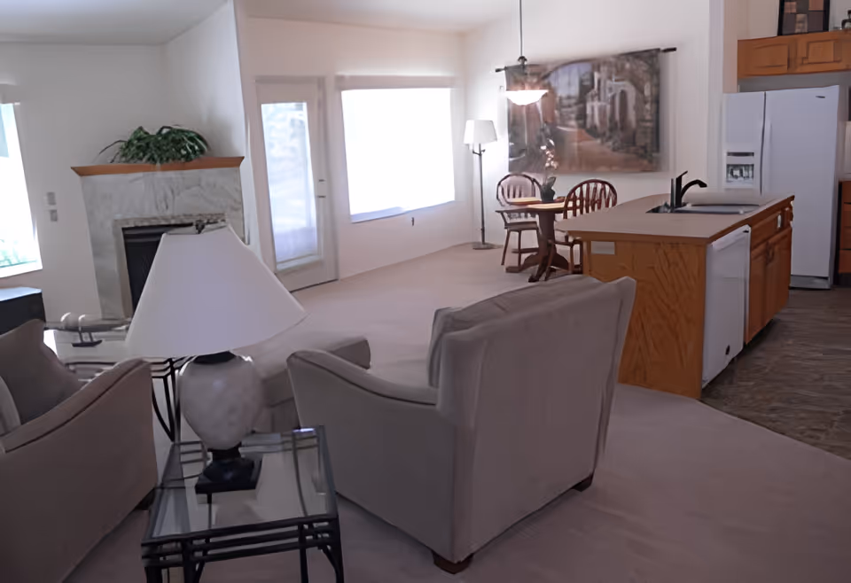 Interior view of a retirement community living space featuring a beige armchair and sofa, a glass-top side table with a white lamp, a stone fireplace with a plant on the mantel, a small round dining table with two wooden chairs, and a kitchen area with wooden cabinets, a white refrigerator, dishwasher, and a sink on the island. Large windows and a glass door allow natural light into the room.