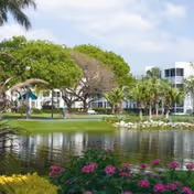 A serene outdoor scene at Five Star Premier Residences of Hollywood featuring a pond with reflections of surrounding trees and shrubs, colorful flowers in the foreground, and a modern white building in the background under a partly cloudy sky.