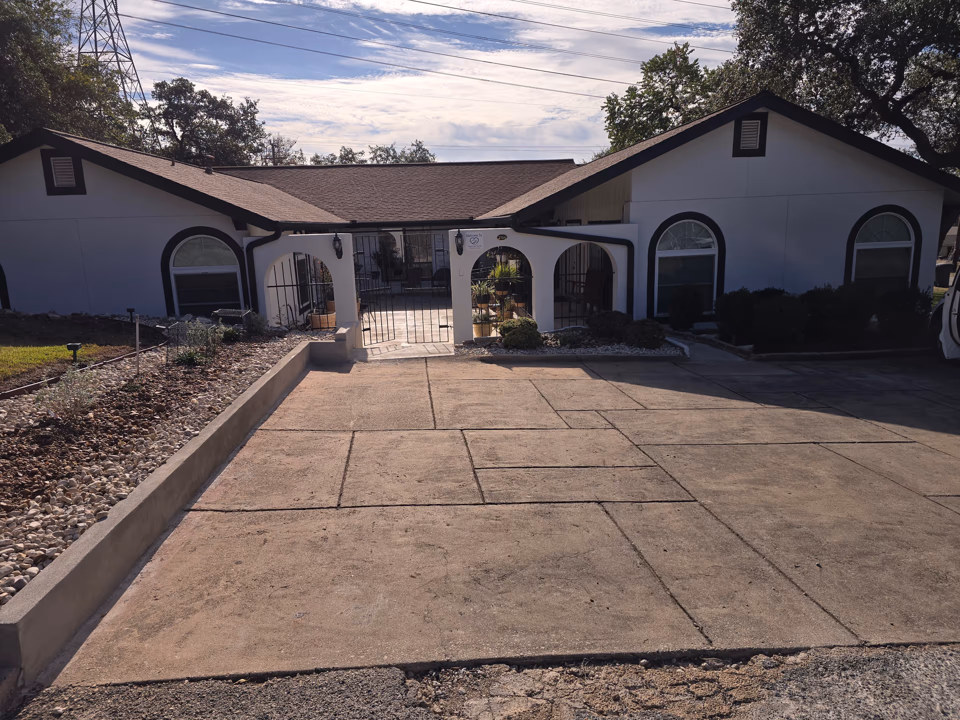 Front exterior view of a single-story building with white walls and dark trim, featuring arched windows and a gated entrance leading to a small patio area with plants. The driveway and landscaped areas with rocks and shrubs are visible in the foreground under a partly cloudy sky.