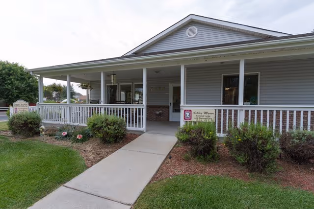 Front exterior view of Ashley Manor Memory Care facility showing a single-story building with a covered porch, white railing, bushes, and a concrete walkway leading to the entrance.