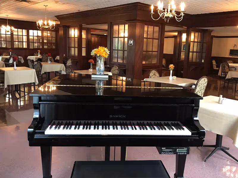 A black grand piano sits in the foreground of a dining room with tables, chandeliers, wood paneling, and floral centerpieces.
