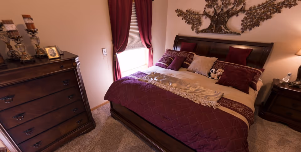A cozy bedroom with a large wooden bed adorned with burgundy and beige bedding, multiple pillows, and two small stuffed animals. There is a wooden dresser with decorative candle holders and a framed photo on top. A window with burgundy curtains allows natural light into the room. A nightstand with a lamp and a telephone is beside the bed. The walls are light-colored and decorated with a metal tree wall art above the bed.