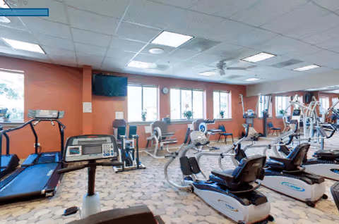 A senior living community fitness room with various exercise equipment including treadmills, stationary bikes, and weight machines. The room has large windows letting in natural light, orange walls, and a wall-mounted TV.