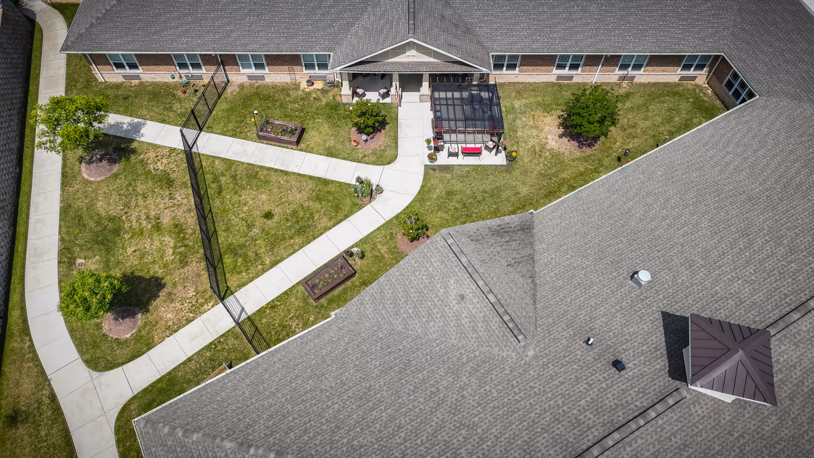 Aerial view of the courtyard area of Chatham Ridge Assisted Living facility showing a building with a gray roof surrounding a grassy area with paved walkways, small trees, and garden beds. There is a covered patio with seating near the center of the courtyard.