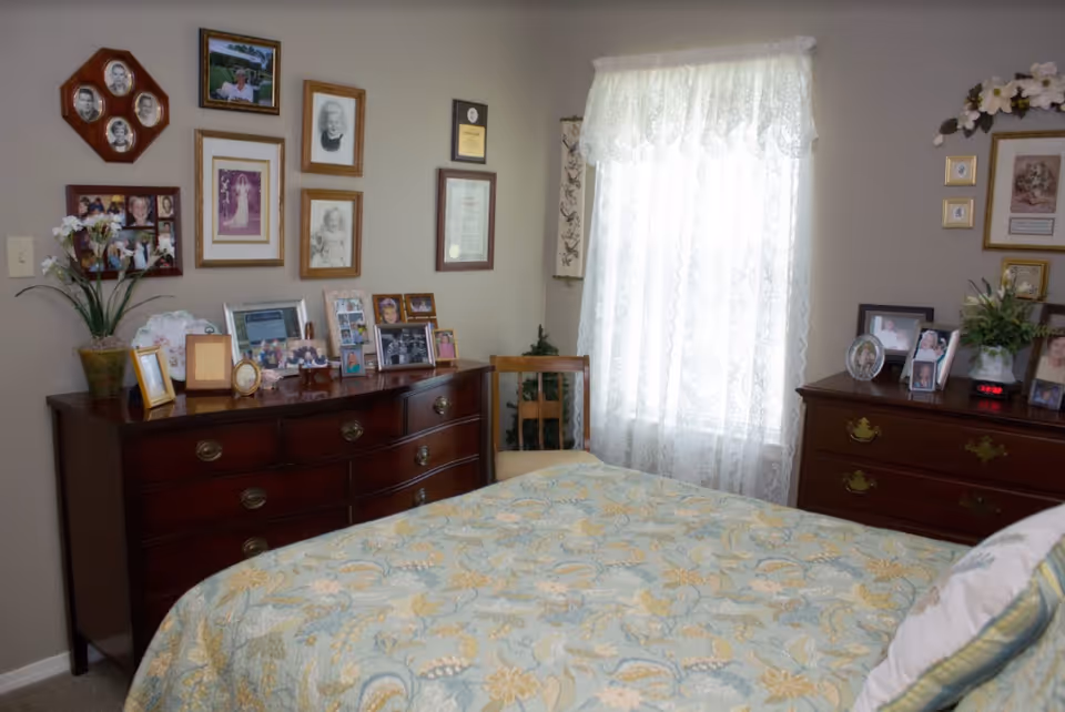 A cozy bedroom with a bed covered in a floral patterned quilt. Two wooden dressers are against the walls, each adorned with numerous framed photographs and decorative items. A wooden chair is placed near a window with sheer lace curtains, allowing soft natural light to fill the room. The walls are decorated with more framed pictures and certificates.