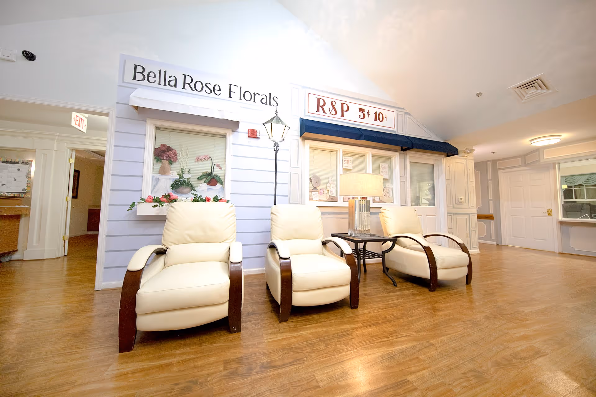 Interior view of a senior living facility lounge area with three cream-colored recliner chairs arranged around a small table with a lamp. The wall behind features decorative storefront-style facades labeled 'Bella Rose Florals' and 'R&P 5¢ 10¢'. The floor is wooden, and the space is well-lit with a clean and welcoming atmosphere.