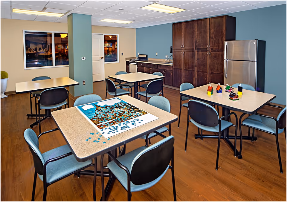 A communal room with several tables and chairs, a kitchenette with dark wood cabinets, a stainless steel refrigerator, and a stove. One table has a partially completed jigsaw puzzle, and another table has small colorful decorative items. The room has wood flooring and walls painted in beige and blue tones.