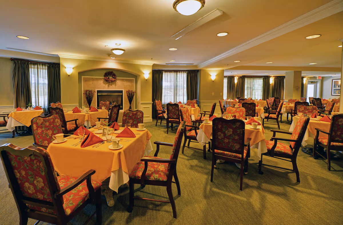 A spacious dining room with multiple tables covered in peach-colored tablecloths and set with cups, glasses, and red folded napkins. The chairs have wooden frames with floral upholstery. The room is warmly lit with ceiling lights and wall sconces, and there are large windows with green curtains. A fireplace with decorative vases and a wreath is visible on one wall.