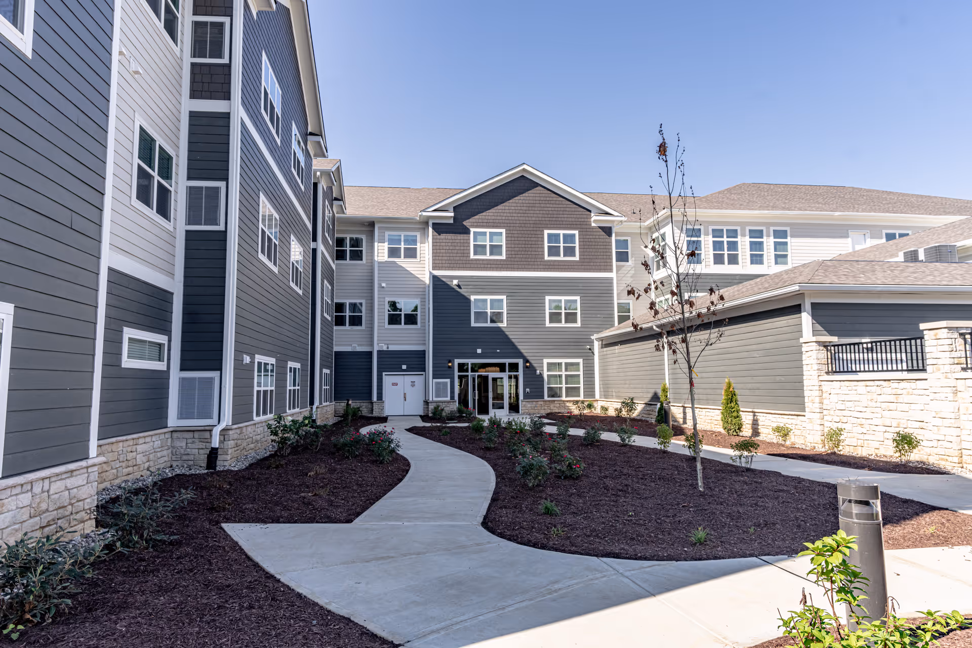 Exterior view of a modern multi-story senior living facility building with gray siding and stone accents. A curved concrete walkway leads through landscaped grounds with small plants and a young tree under a clear blue sky.